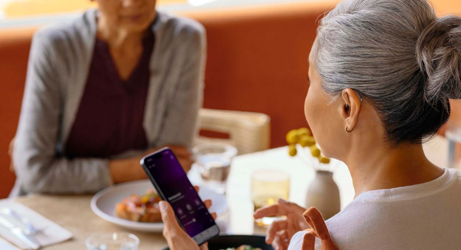 A woman wearing hearing aids using her phone app while talking to another woman at a restaurant